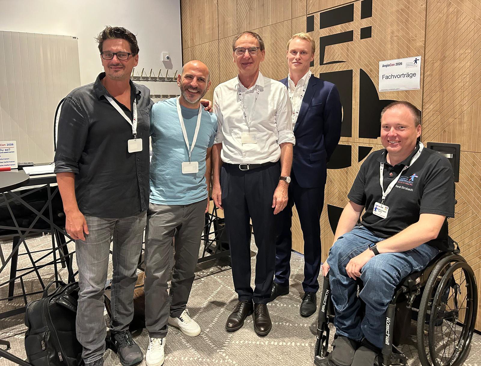 Cinq hommes portant un badge du congrès, dont un en fauteuil roulant, posent côte à côte devant une paroi en bois portant l'écriteau « Fachvorträge ».